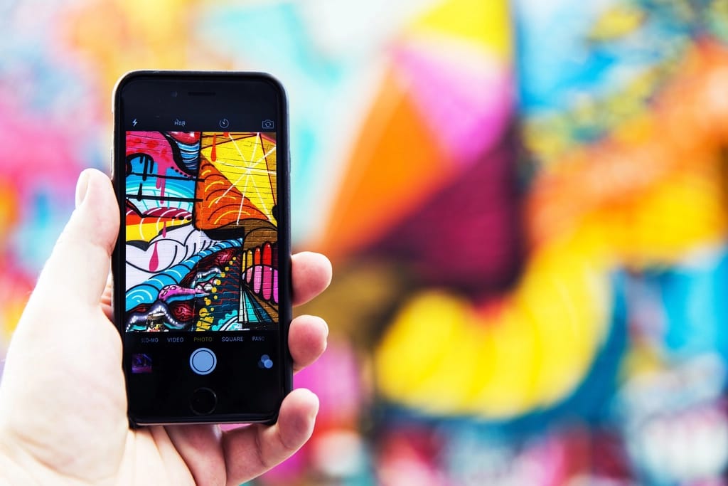 A hand holds up a phone framing a large, colourful street-art mural; the artwork appears crisp on the screen and blurred behind.