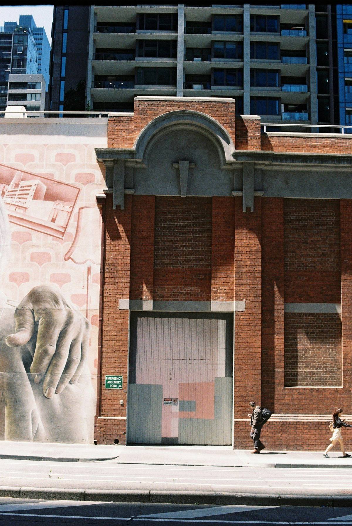 A large mural on the side of a brick building on a city street, two people walking past it on the pavement.