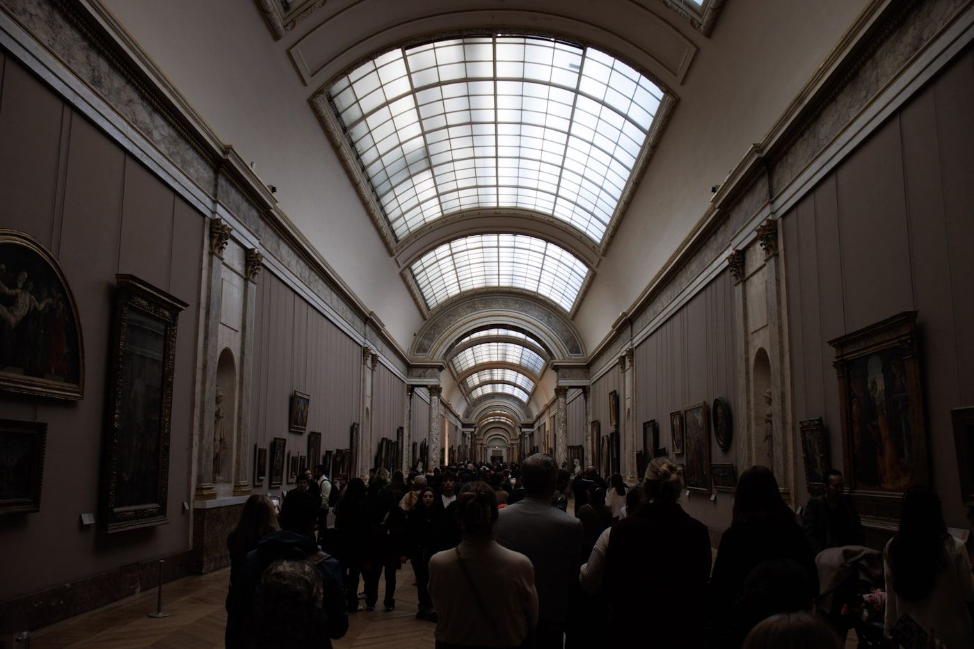 A long, high-ceilinged museum gallery lined with framed paintings, crowded with visitors walking through it.