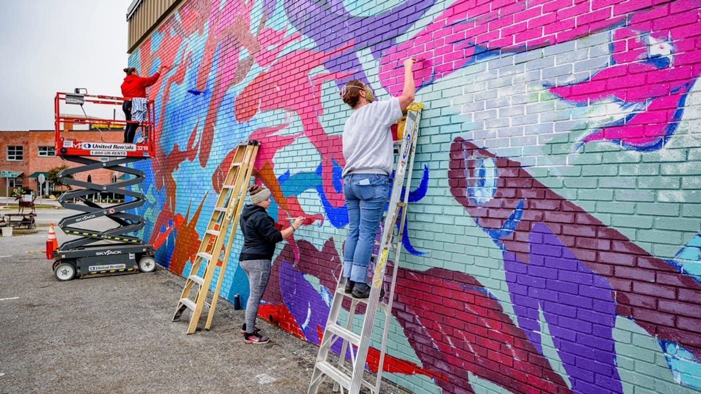 Three people on ladders and a lift painting a large, colourful mural across the brick side of a downtown building.