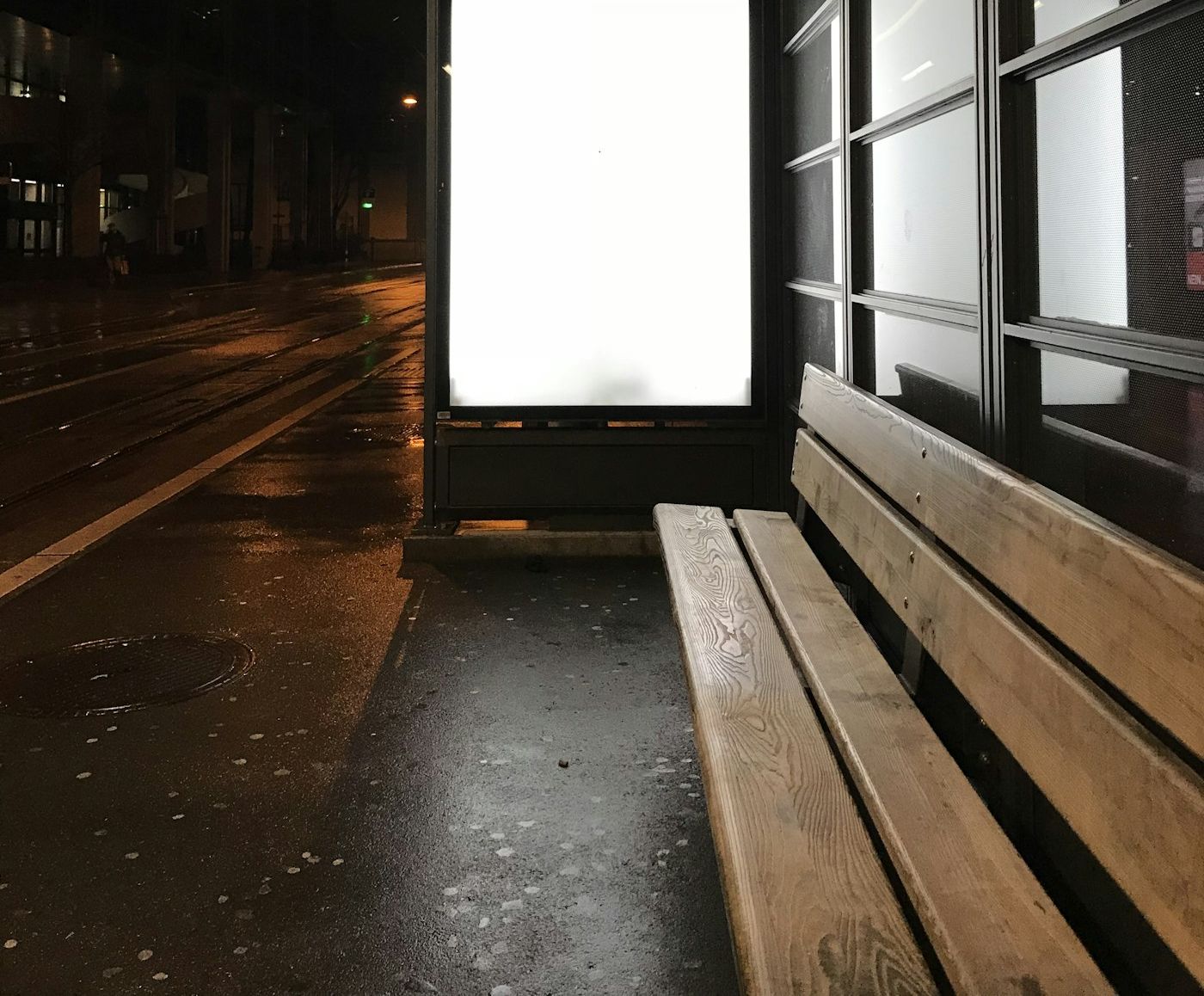 An empty illuminated advertising panel at a transit shelter on a wet street at night, the pavement reflecting its light.