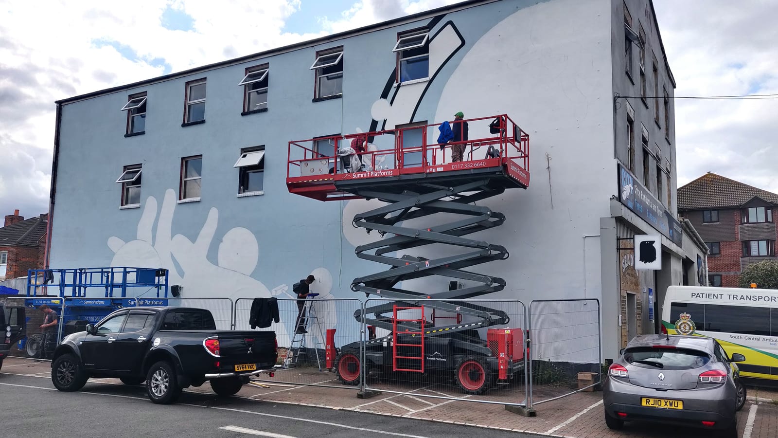 A mural half-painted on the side of a three-storey building. Two artists work from a raised scissor lift and a step ladder, with a second lift, safety barriers and vehicles parked at the base of the wall.