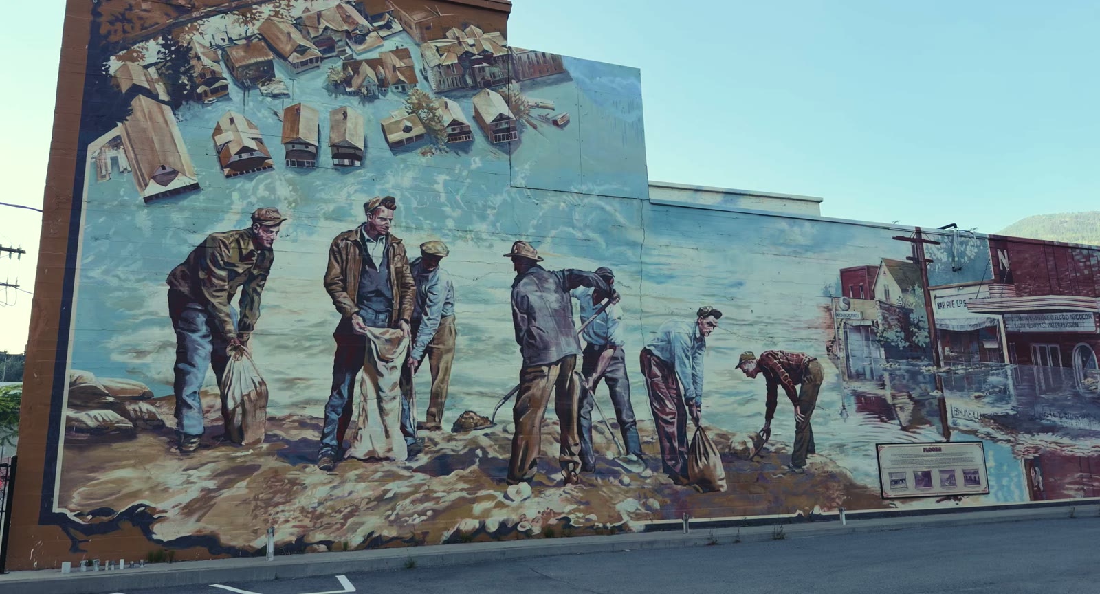 The Flood mural on a blank hotel wall in Trail, BC — a painted crew filling sandbags along the riverbank, with an empty parking lot in front of it and nobody stopping to look.